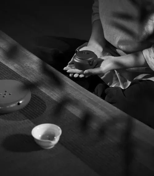 Fotografía en blanco y negro de una persona que sostiene un pequeño objeto tetera junto a una mesa de té