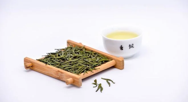 Flat green tea leaves in a bamboo scoop beside a cup