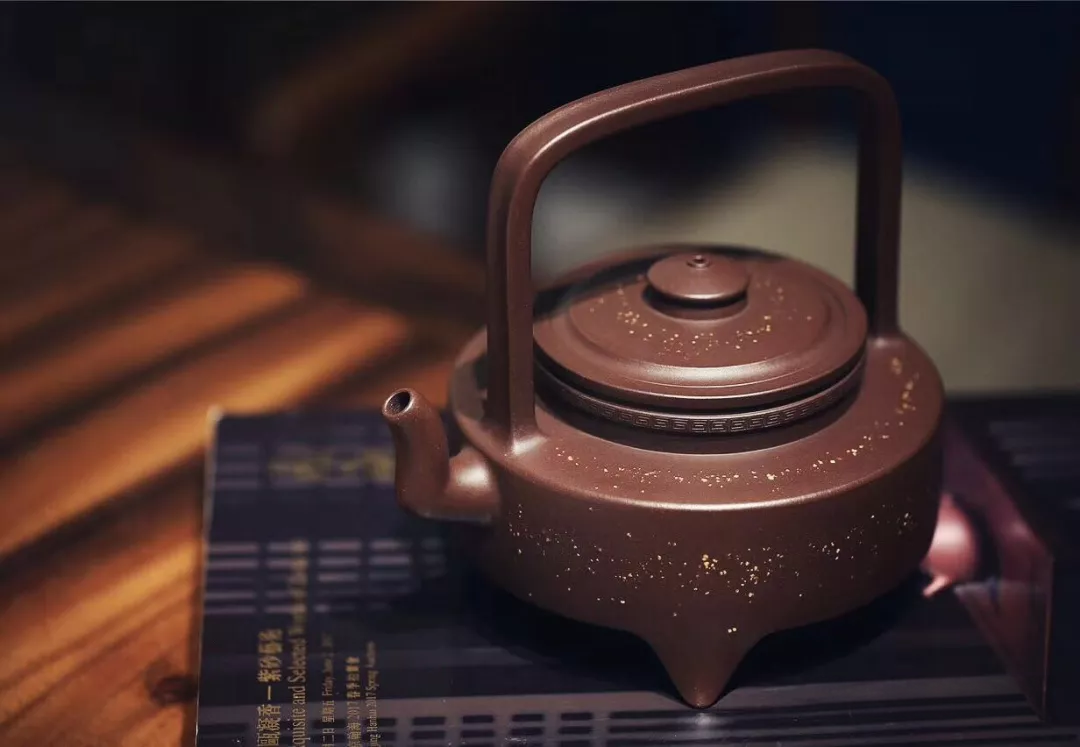 A three-footed clay teapot with a tall overhead handle and pale decorative speckles
