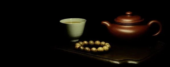 A brown clay teapot and pale cup on a dark tea tray with a strand of beads